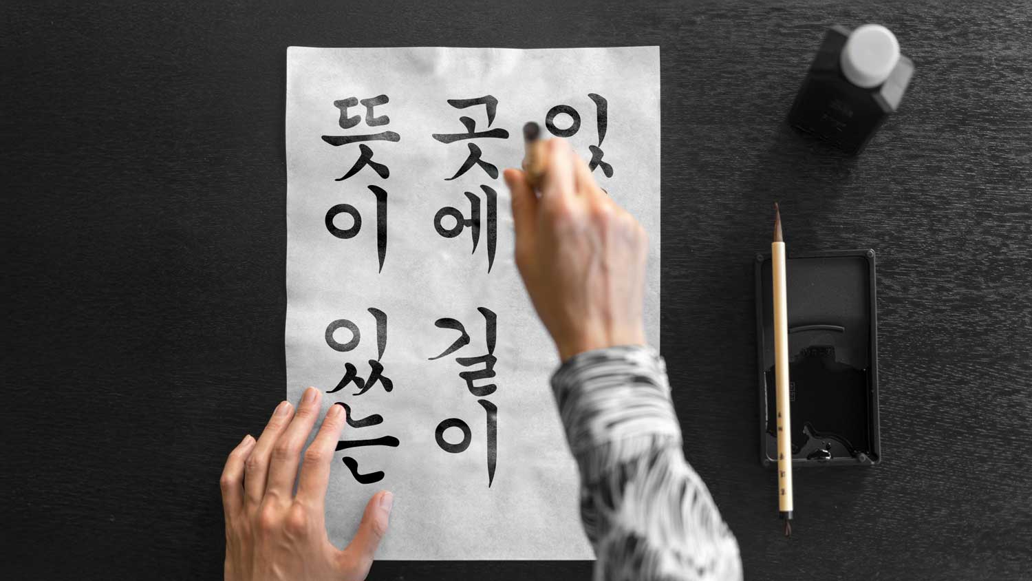 Gros plan sur les main d'une personne tenant un pinceau au dessus d'une feuille comportant une calligraphie  coréenne traditionnelle