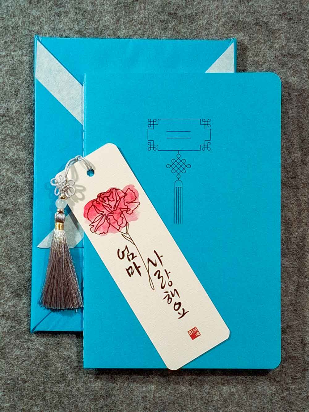 Marque page calligraphié "je t'aime maman" en coréen, avec un nœud coréen, un carnet décoré et un paquet cadeau plié en forme de hanbok. Le tout dans les tons bleu