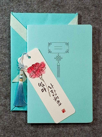Marque page calligraphié "je t'aime maman" en coréen, avec un nœud coréen, un carnet décoré et un paquet cadeau plié en forme de hanbok. Le tout dans les tons bleu pastel