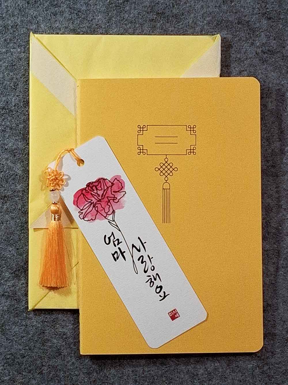Marque page calligraphié "je t'aime maman" en coréen, avec un nœud coréen, un carnet décoré et un paquet cadeau plié en forme de hanbok. Le tout dans les tons jaune