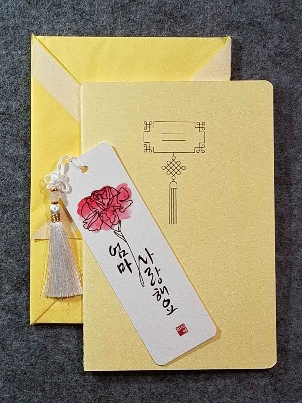 Marque page calligraphié "je t'aime maman" en coréen, avec un nœud coréen, un carnet décoré et un paquet cadeau plié en forme de hanbok. Le tout dans les tons jaune pastel