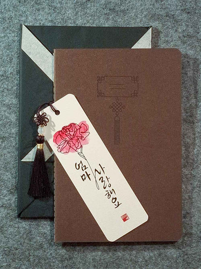 Marque page calligraphié "je t'aime maman" en coréen, avec un nœud coréen, un carnet décoré et un paquet cadeau plié en forme de hanbok. Le tout dans les tons marron