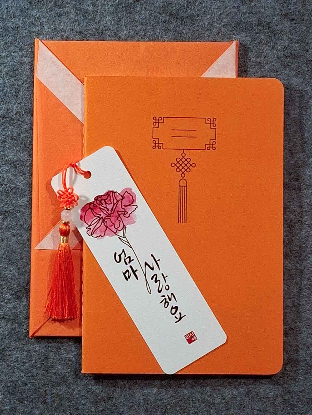 Marque page calligraphié "je t'aime maman" en coréen, avec un nœud coréen, un carnet décoré et un paquet cadeau plié en forme de hanbok. Le tout dans les tons orange