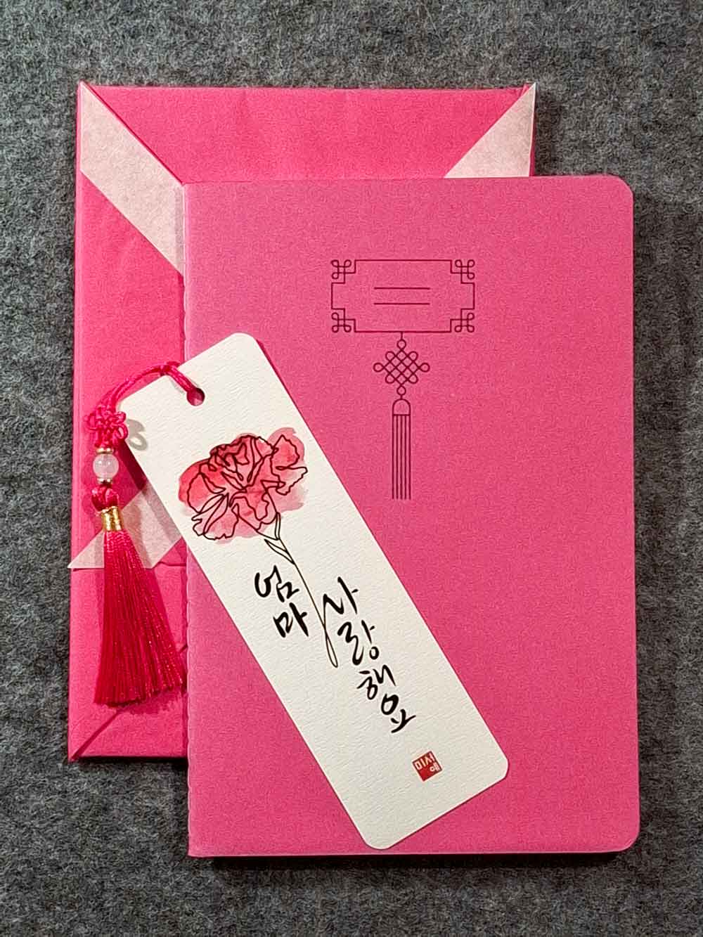 Marque page calligraphié "je t'aime maman" en coréen, avec un nœud coréen, un carnet décoré et un paquet cadeau plié en forme de hanbok. Le tout dans les tons rose carmin