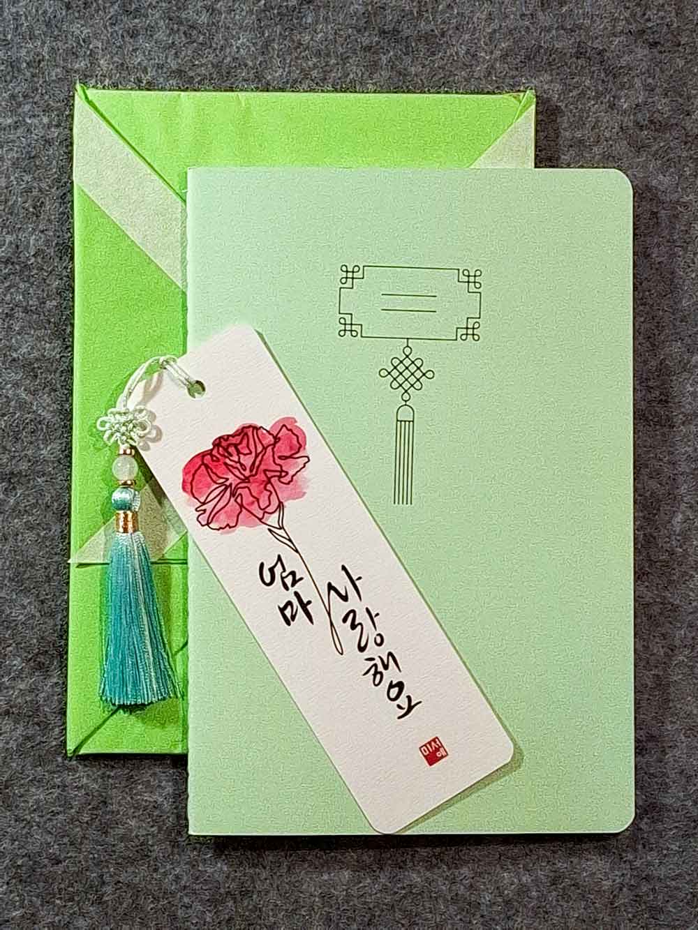 Marque page calligraphié "je t'aime maman" en coréen, avec un nœud coréen, un carnet décoré et un paquet cadeau plié en forme de hanbok. Le tout dans les tons vert amande