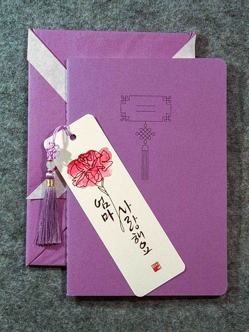 Marque page calligraphié "je t'aime maman" en coréen, avec un nœud coréen, un carnet décoré et un paquet cadeau plié en forme de hanbok. Le tout dans les tons lavande