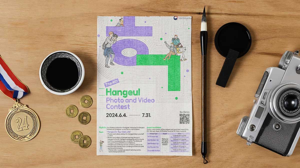 Affiche du concours photo et vidéo sur le thème du Hangeul, l'affiche est posé sur une table en bois avec autour un pinceau, de l'encre de chine, un appareil photo, une médaille et des pièces de monnaie